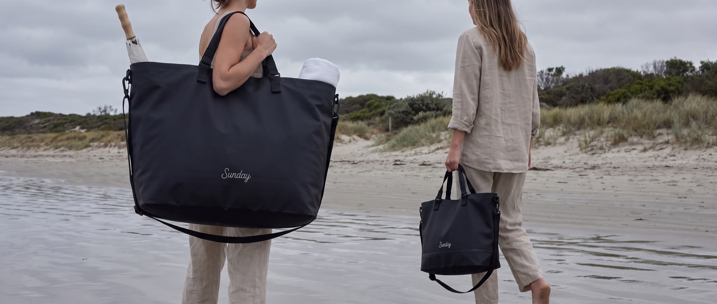Two women walking up off the beach at the end of the day, one with the Sunday Tote XL on her shoulder and one carrying the Sunday Tote
